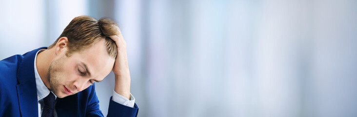 Fototapeta premium Stressed, tired businessman or heaving headache, holding his head, in blue suit, at office. Business fall, job concept. Wide banner composition image with copy space.