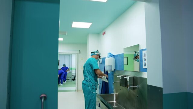 Male mid-aged neurosurgeon wearing device glasses on head stands in the preoperational room. Doctor uses napkins to dry his hands after washing.