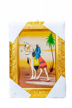 A sand painting with a gold frame, the corners protected by white paper, represents a Saharan rider holding a sword and wearing traditional head scarf on a white camel with clothing and a palm tree.