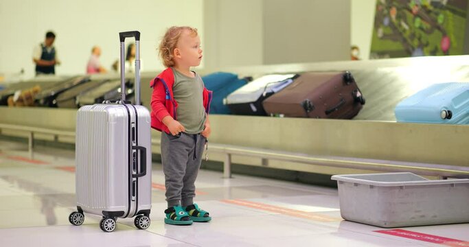 Young boy stand by trolley case at baggage claim hall area of large airport, slow motion full length portrait shot. Suitcases move on conveyor, seen blurred on background
