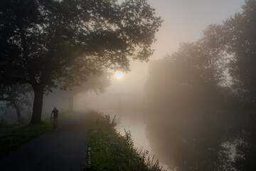 A solitary cyclist embarks on a journey along a riverside path shrouded in mist in this evocative...