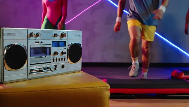 Cropped shot of people performing aerobics steps to music with focus on boombox in studio with neon lights