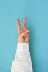 A hand in a white shirt shows the V gesture with two fingers on a blue background - obrazy, fototapety, plakaty