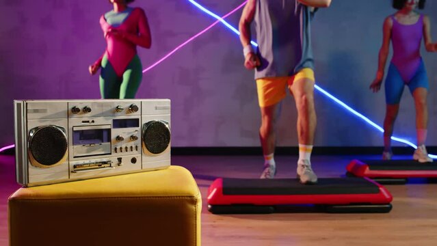 Cropped slowmo shot of unrecognizable friends stepping to boombox music while practicing aerobics in retro outfits in neon studio