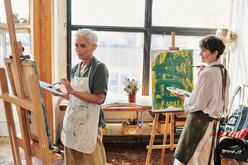 talented mature female friends in aprons painting on easels in modern art workshop, creative hobby