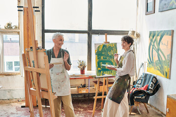 happy middle aged women in aprons painting on easels in modern art workshop, creative leisure