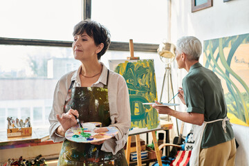 Fototapeta na wymiar inspired mature woman holding palette while female friend painting in art workshop, creative leisure