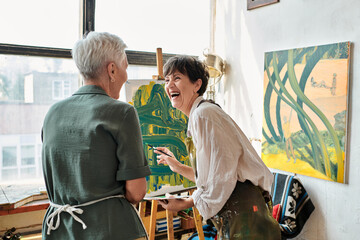 Obraz na płótnie Canvas excited mature woman laughing near easel and female friend in craft workshop, creative hobby