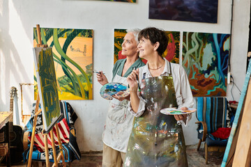 happy stylish mature women in aprons looking away in modern art workshop, friendship and creativity