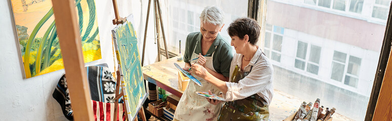 smiling middle aged women mixing colors on palettes near easels in art studio, horizontal banner