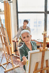 inspired mature woman in apron painting on easel at master class in art workshop, creative hobby