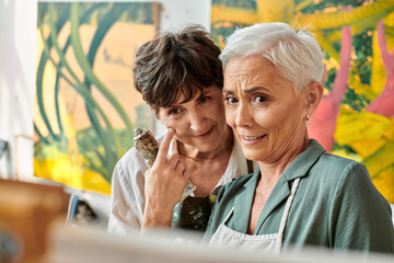 positive and pensive mature women looking at easel during painting master class in art workshop