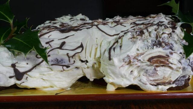 Closeup dolly shot of a yule log dessert (also called a chocolate roulade or a Bûche de Noël). Sponge cake covered in thick white cream and splashes of brown chocolate, with sprigs of holly.