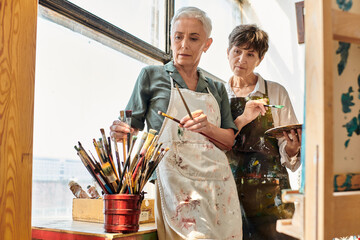 skilled female artists choosing  paintbrushes near mature woman during master class in art workshop