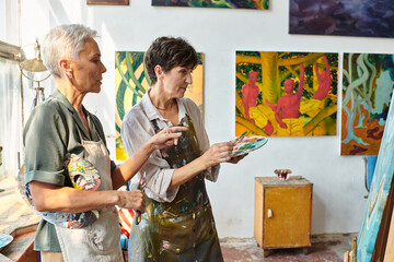 skilled female artist pointing at easel near mature woman with palette during master class in studio