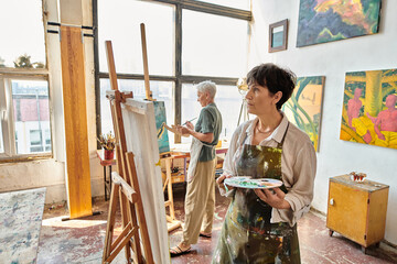 thoughtful mature woman in apron looking at easel near female friend in modern spacious art workshop