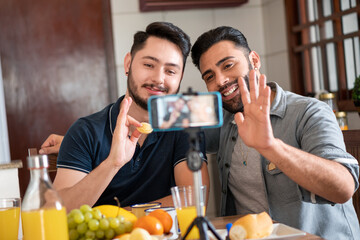 happy gay couple smiling and waving mobile phone on video call in kitchen table