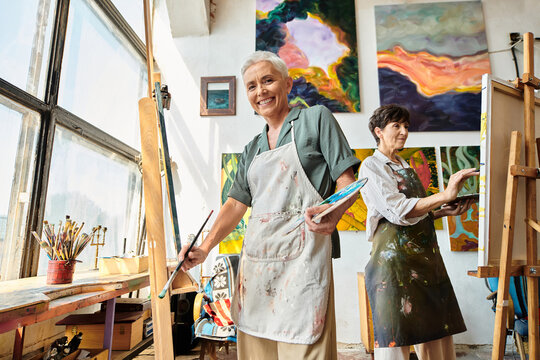 overjoyed mature woman in apron looking at camera in modern spacious workshop, artistic hobby