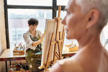 skilled female artist painting portrait on blurred mature model in craft workshop, inspiration