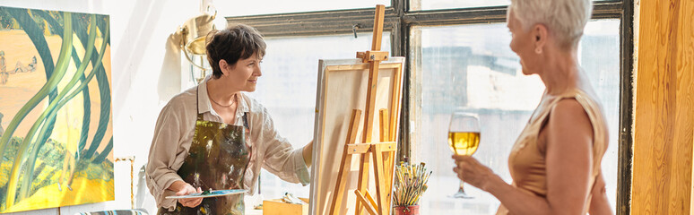 talented female artist painting elegant mature model with wine glass in art studio, banner