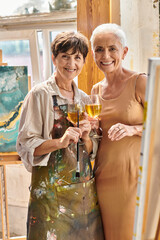 delighted female painter and stylish mature model with wine glasses looking at camera in workshop