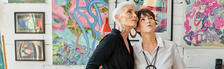 stylish middle aged lesbian couple looking at creative paintings in modern art workshop, banner