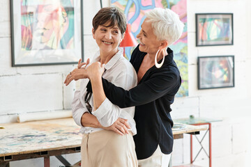cheerful and trendy mature woman embracing lesbian partner in modern art workshop, happiness