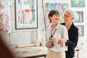 joyful lesbian artists embracing and looking at each other in modern art workshop, bonding