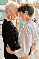 stylish mature lesbian artists standing face to face and smiling with closed eyes in art workshop