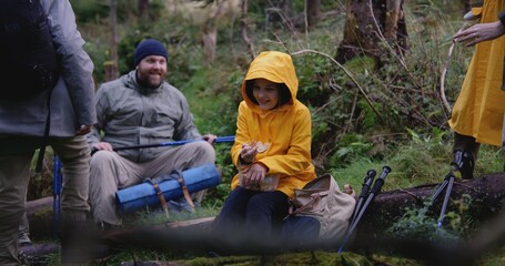 Young hiker sits on log and takes out snacks from food bowl. Group of outdoor enthusiasts rest in camp after long walking trek in the mountains. Tourism and outdoor exploration concept. Slow motion.