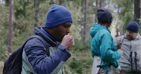 African American tourist keeps cup in hand and shares hot tea with female traveler. Group of multiethnic hiking buddies stopped to rest in forest camping. Nature discovery and tourism. Slow motion.