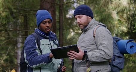 Two multiethnic tourists discuss trail way using tablet computer in forest. Outdoor enthusiasts with backpacks and trekking poles stop to rest during hike. Nature discovery and tourism. Slow motion.