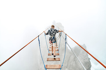 Courageous boy walks along rope bridge in mountain park on foggy day. Little tourist enjoys extreme...