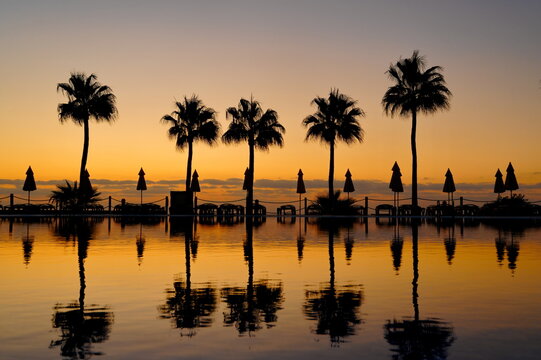 Sonnenaufgang am Pool mit Spiegelung auf dem Wasser, Palmen und Sonnenschirme im Hintergrund