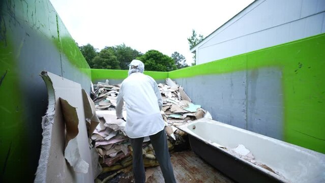 Camera following a female construction worker wearing a respirator through a demolition site as she carries pieces of wallboard to a dumpster.