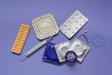 Contraceptive pills, condoms, intrauterine device and thermometer on violet background, flat lay....