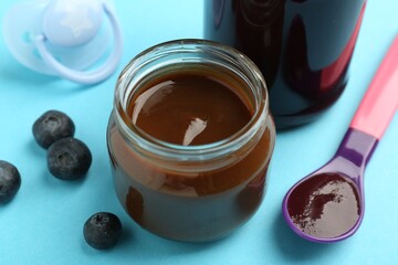 Jars with healthy baby food, spoon, blueberries and pacifier on light blue background, closeup
