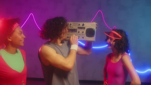 Medium shot of male trainer loading cassette into retro tape recorder during dancing or aerobics class with female friends dancing to music in neon studio
