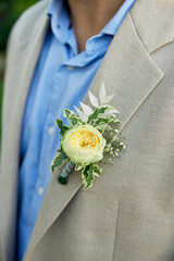 Close-up captures groom in light grey suit, blue shirt, adorned with elegant boutonniere pinned to lapel. Boutonniere features cream-colored rose, surrounded by green leaves, smaller white flowers