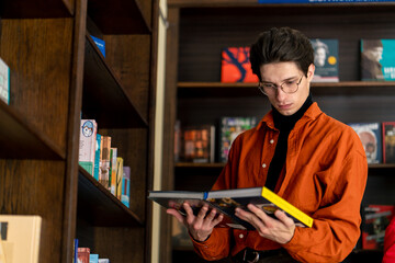 young guy in glasses standing near bookshelves holding two books in hands choosing what to read or...