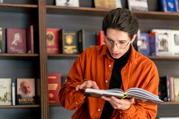 young guy in glasses stands near the bookshelves, holds reads a book enthusiastically and marvels...