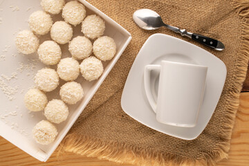Several candies with nut and coconut flakes in paper box with ceramic saucer and cup on jute napkin, macro, top view.