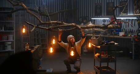 Female carpenter completes wood processing, looks at finished design product from tree branch with light bulbs. Woman artisan woodworking in modern workshop. Handcrafted and entrepreneurship concept.