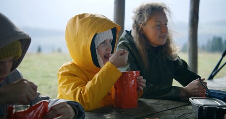 Young girl sits at the table and eats hiking food from pouch during trek. Caucasian woman talks...