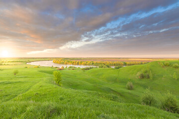 Fototapeta na wymiar A river against a backdrop of green fields, a Bright spring landscape against the backdrop of the sunset sky, green hills illuminated by the setting sun