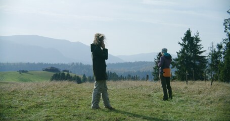 Female tourists stand on the top of the hill and use their mobile phones. One woman tries to catch signal, another talks by phone. Group of hikers during trip or trek in the mountains in autumn.