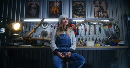 Female carpenter, artisan smiles and looks at camera sitting in modern stylish workshop. Wooden product from tree branch and board with professional tools in background. Handmade and small business.