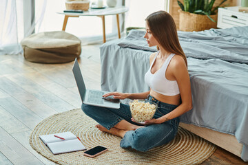 attractive woman in comfy homewear sitting on floor and working at her laptop with popcorn in hand
