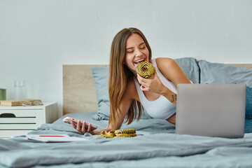 joyous beautiful woman with long hair holding phone and enjoying donut while working from home