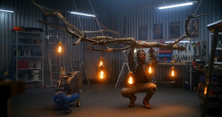 African American carpenter ties with rope design wood product from tree branch with light bulbs. Artisan or designer woodworking in modern garage or workshop. Hand craft and entrepreneurship concept.